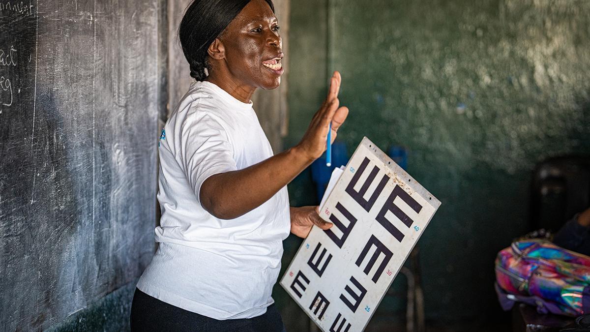 Mitarbeiterin von GoodVision Malawi erklärt vor der Klasse den Ablauf des Sehtests und die Sehprobentafel